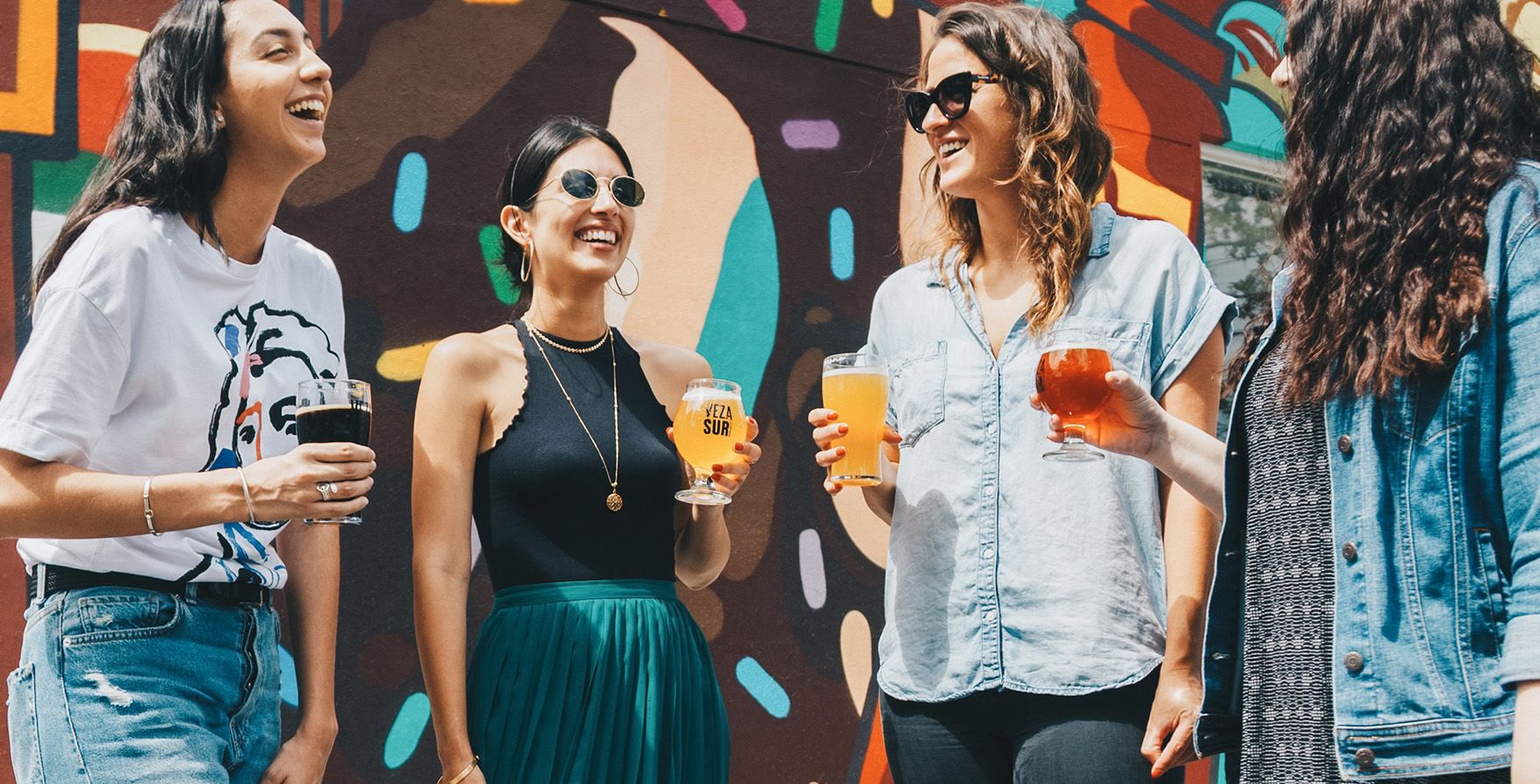 cuatro amigas riendo con bebidas delante de un mural